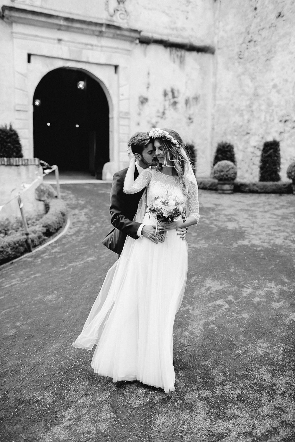 Black and white photo of bride and groom embracing in front of a historic stone archway entrance.