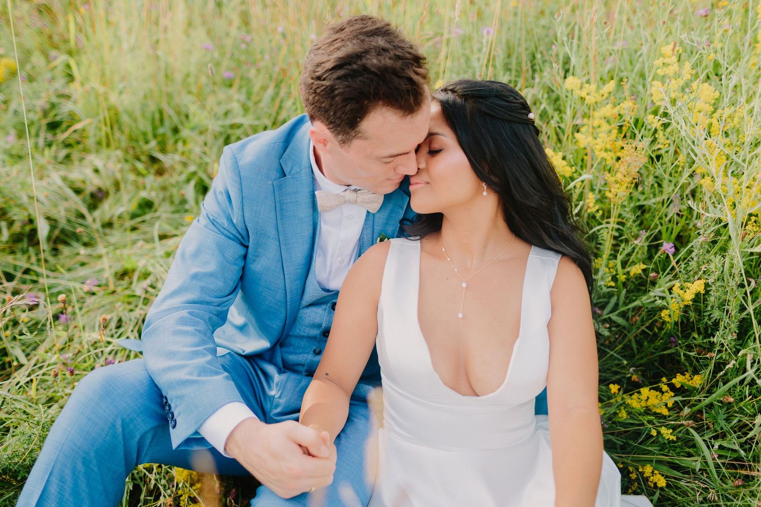 Bride and groom sitting closely in a wildflower field, groom in blue suit touching foreheads romantically.