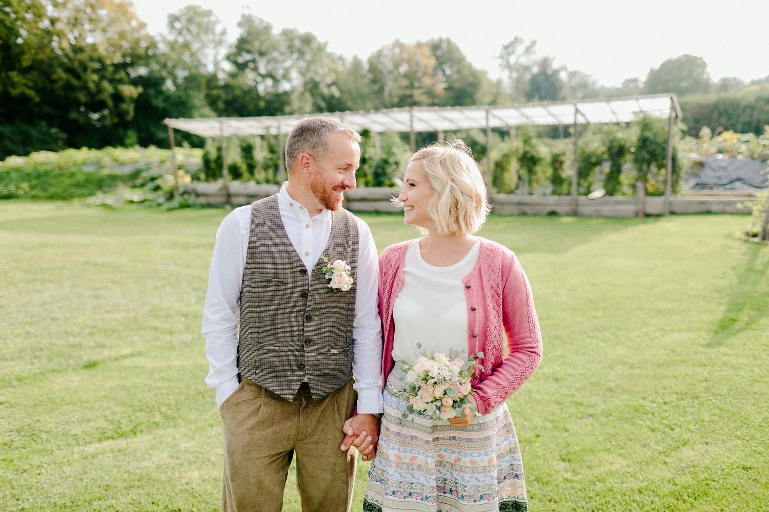Couple holding hands and smiling at each other in a lush green garden during their outdoor wedding celebration.