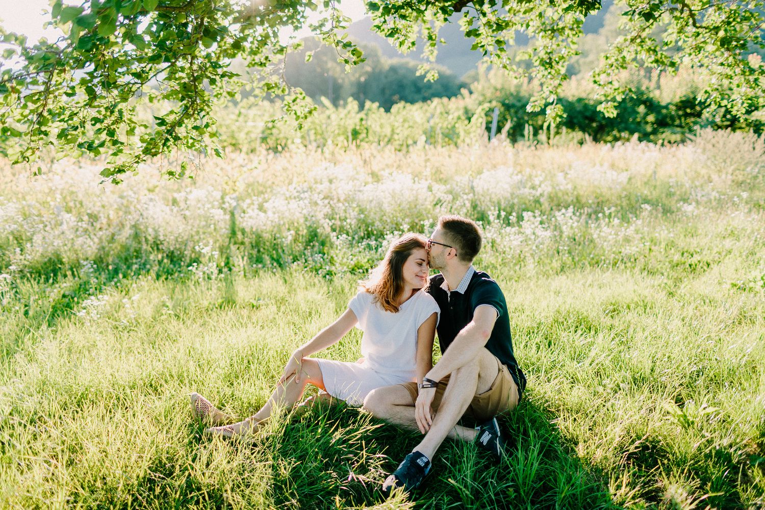 A couple sitting together in a sunny green meadow beneath leafy tree branches.