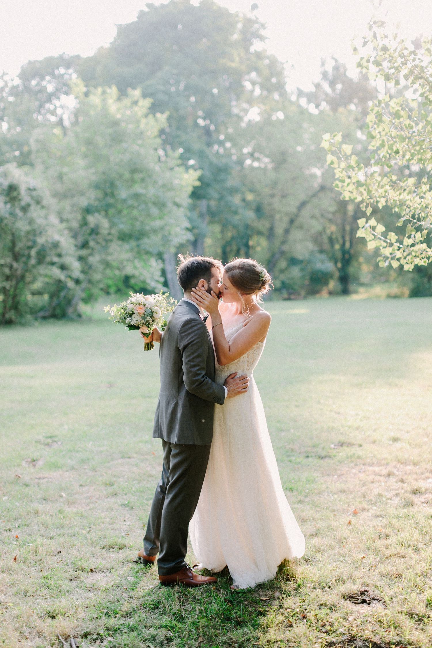 Newlyweds share a romantic kiss in a sunlit green meadow, groom holding bridal bouquet in natural setting.