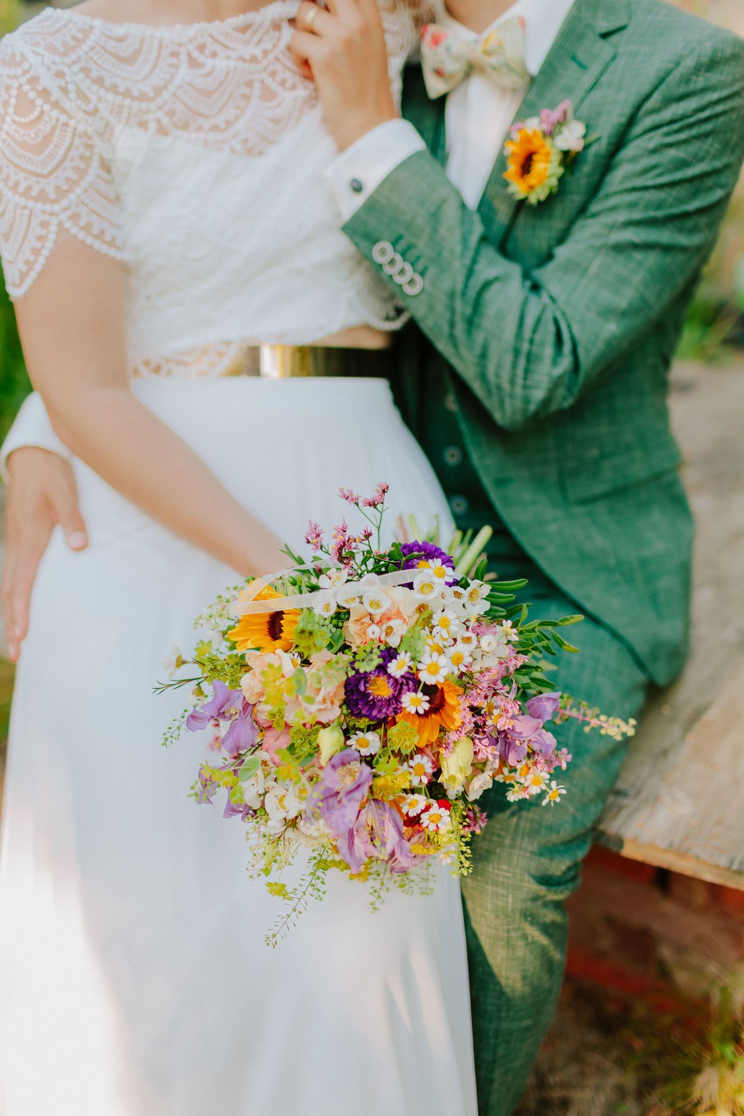 Close-up of vibrant wildflower wedding bouquet with sunflowers held by bride in lace dress beside groom in teal suit.