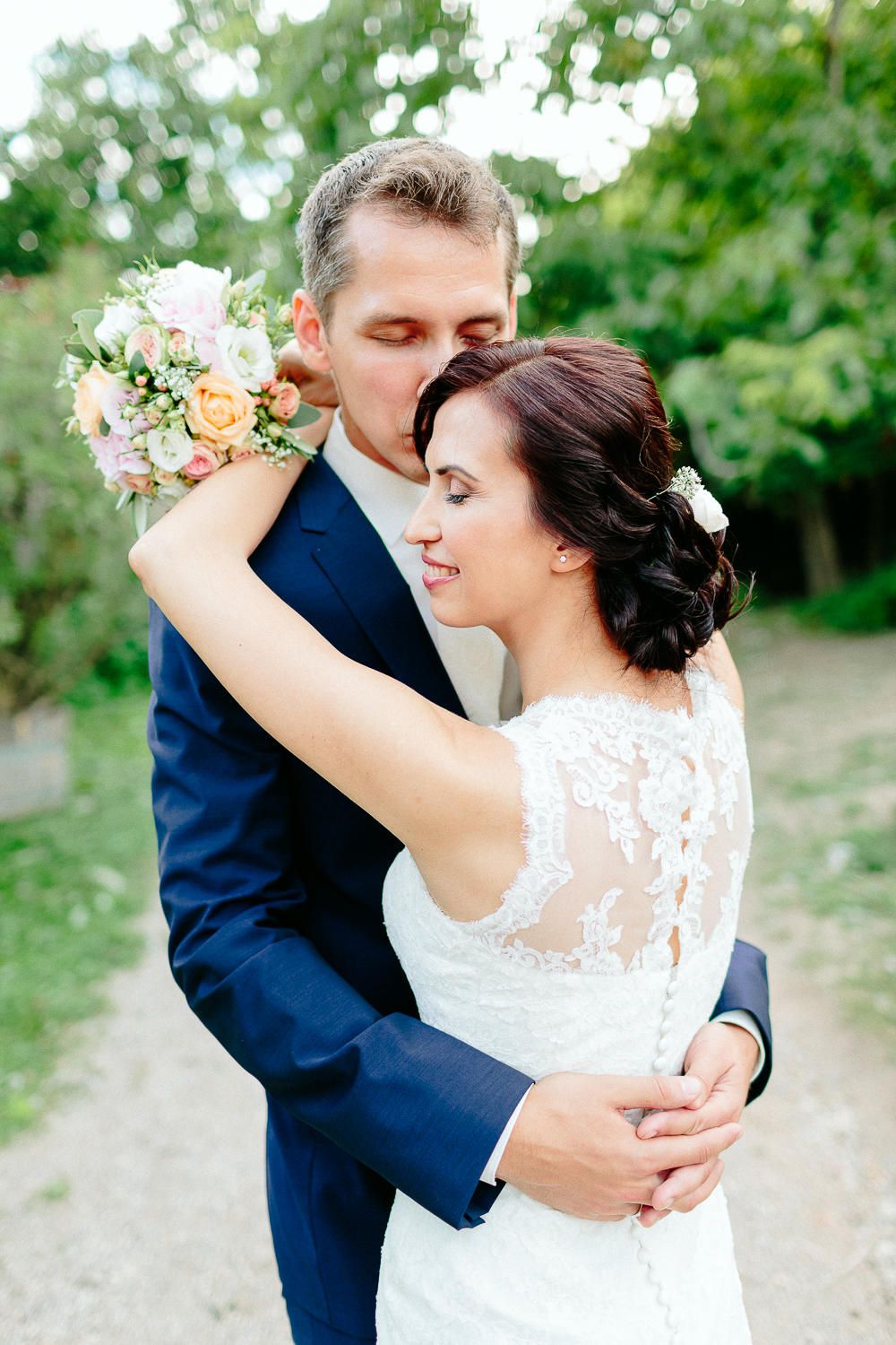 Groom in navy suit kissing bride on head while embracing her in a lush garden setting.