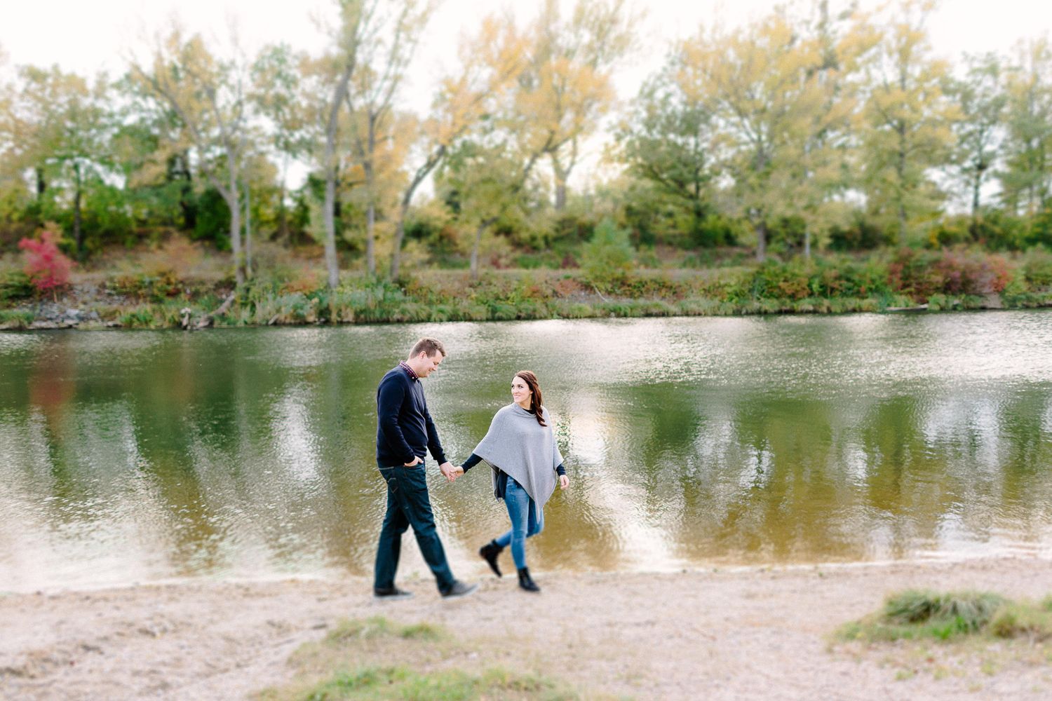 Couple holding hands and walking along a sandy riverbank surrounded by colorful autumn trees.