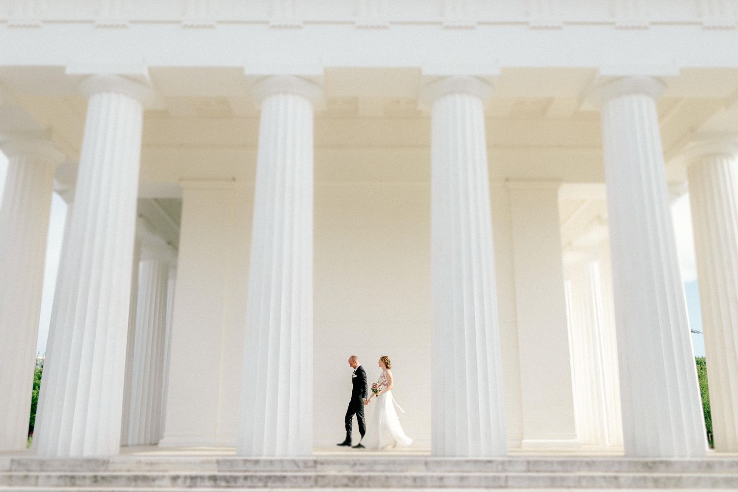 Bride and groom walk between grand white columns at a neoclassical monument on their wedding day.