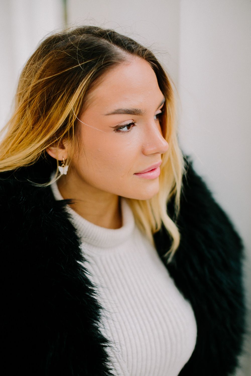 Young woman with blonde ombre hair wearing a black fur coat and white ribbed sweater posing indoors.