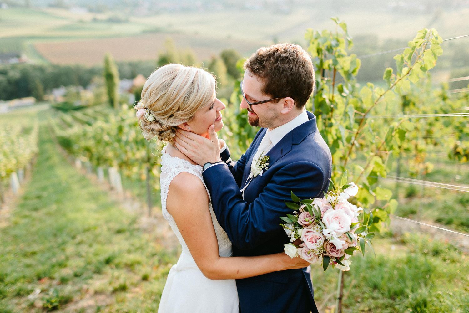 Bride and groom embrace romantically in scenic vineyard with rolling hills in background.