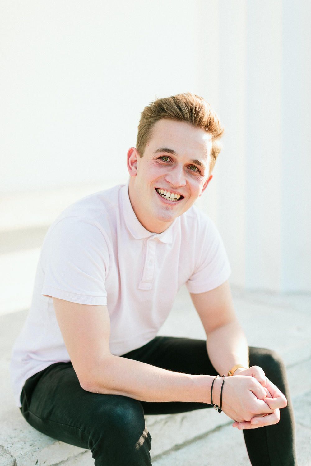 Smiling young man in white polo shirt sitting on steps in bright outdoor setting.
