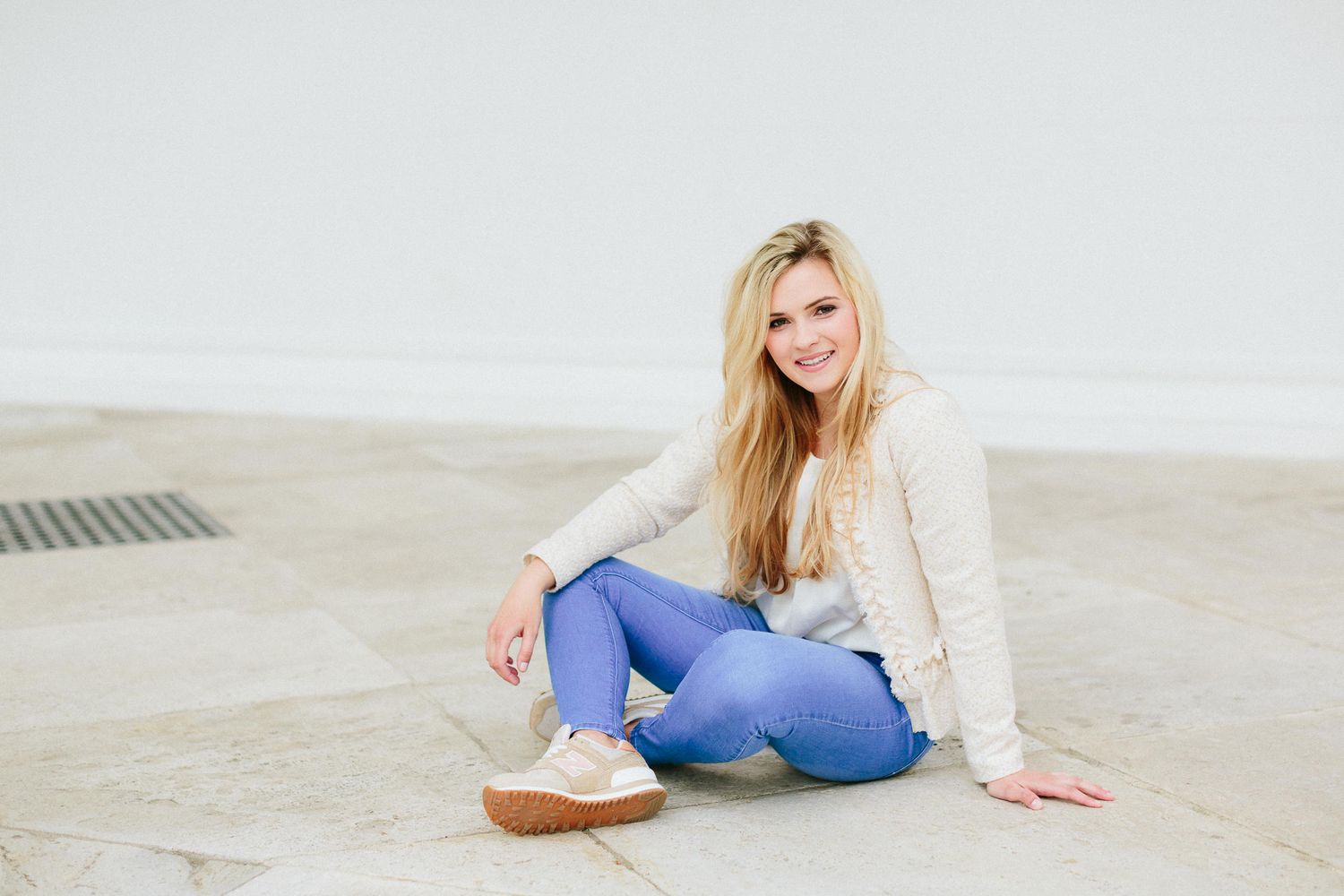 Young blonde woman sitting on stone floor smiling, wearing blue jeans and white sweater.