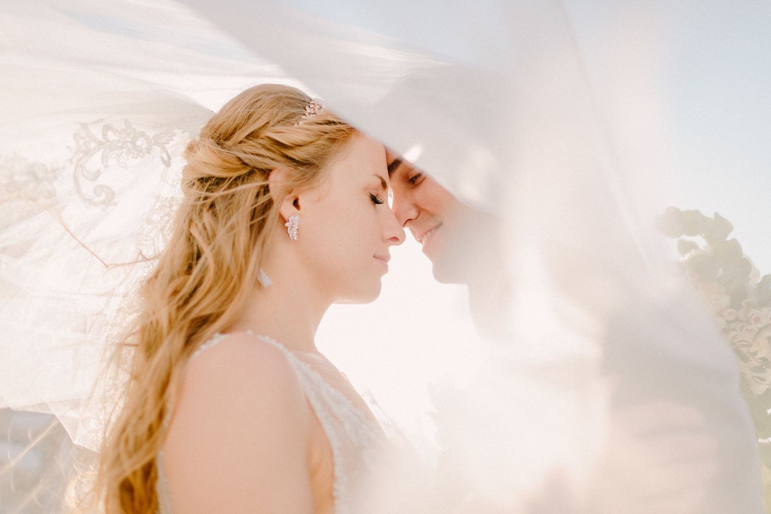 Bride and groom share an intimate moment under flowing white veil in soft dreamy backlit wedding portrait.