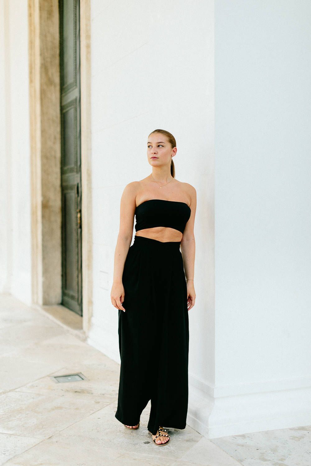 Elegant woman in black strapless crop top and wide-leg pants standing against white wall.