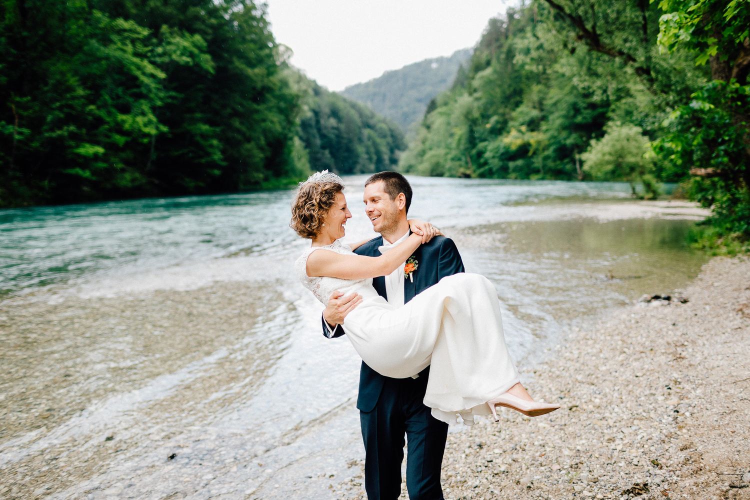 Groom carrying smiling bride in white gown along rocky riverbank with lush green mountain forest backdrop.