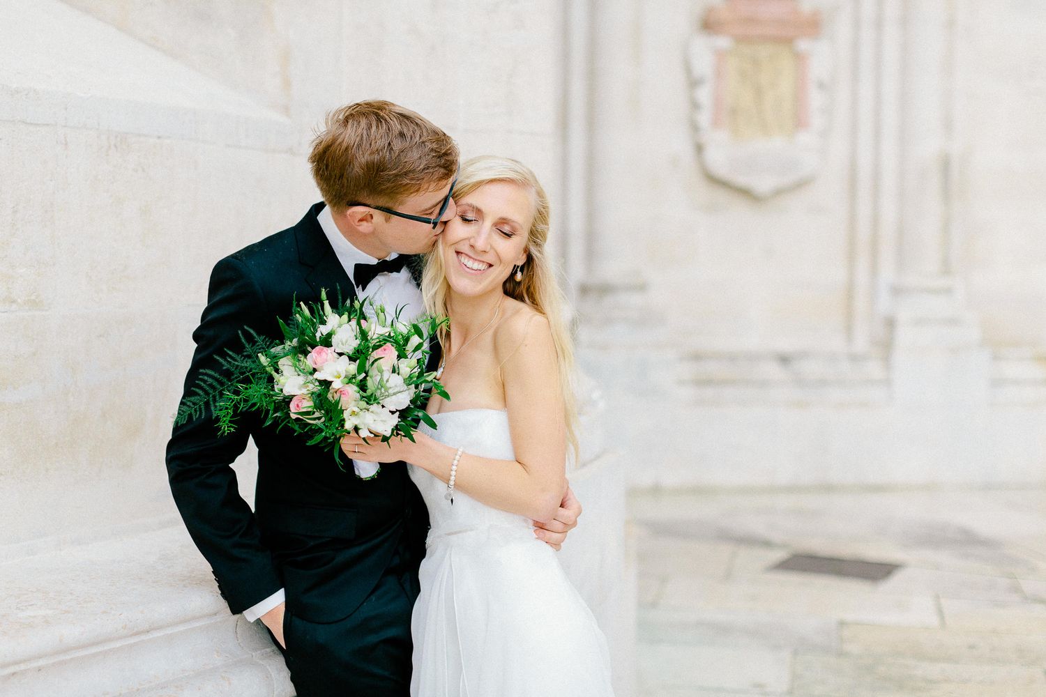 Groom in black tuxedo kissing blonde bride holding lush bouquet against elegant white stone architecture.