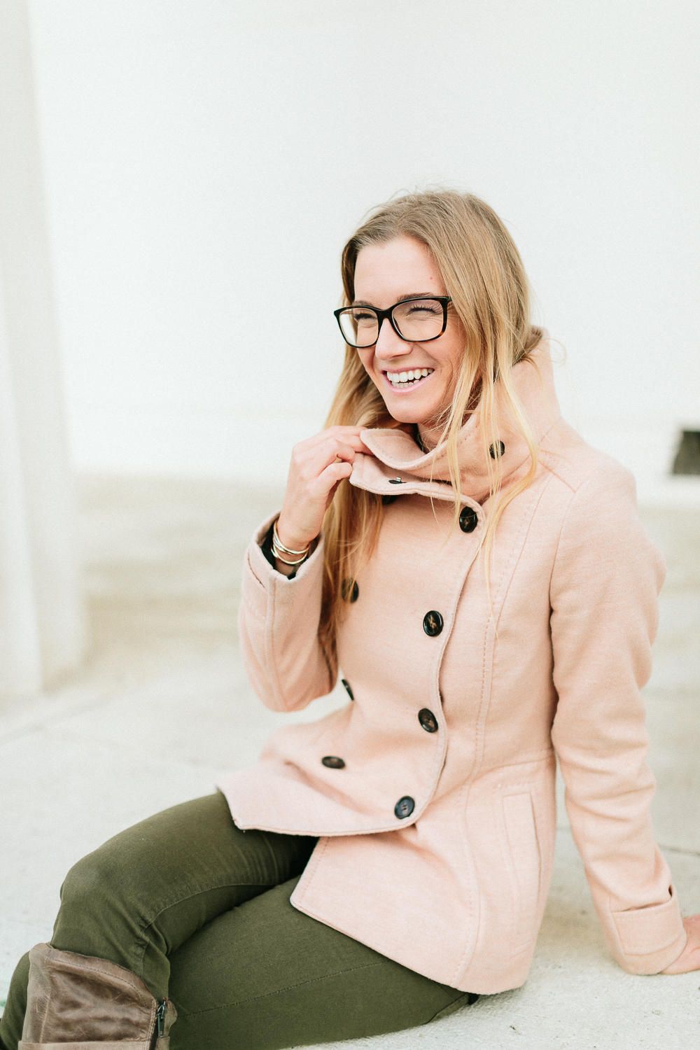 Laughing blonde woman in pink peacoat and glasses sitting outdoors on stone steps.