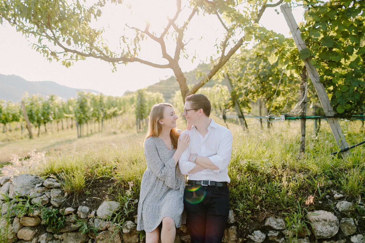 Couple sharing a gentle kiss near a stone wall in a sunny vineyard garden.