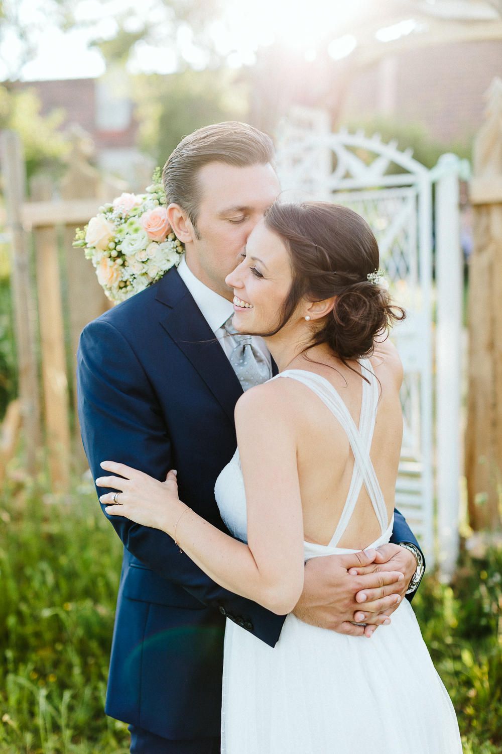 Groom in navy suit embraces smiling bride in backless white dress in a sunlit garden setting.