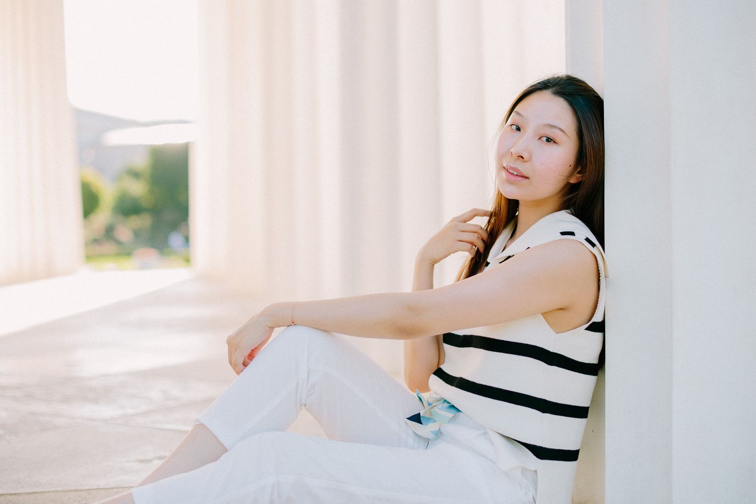 Young Asian woman in a striped top and white pants sitting against a white pillar in bright sunlight.