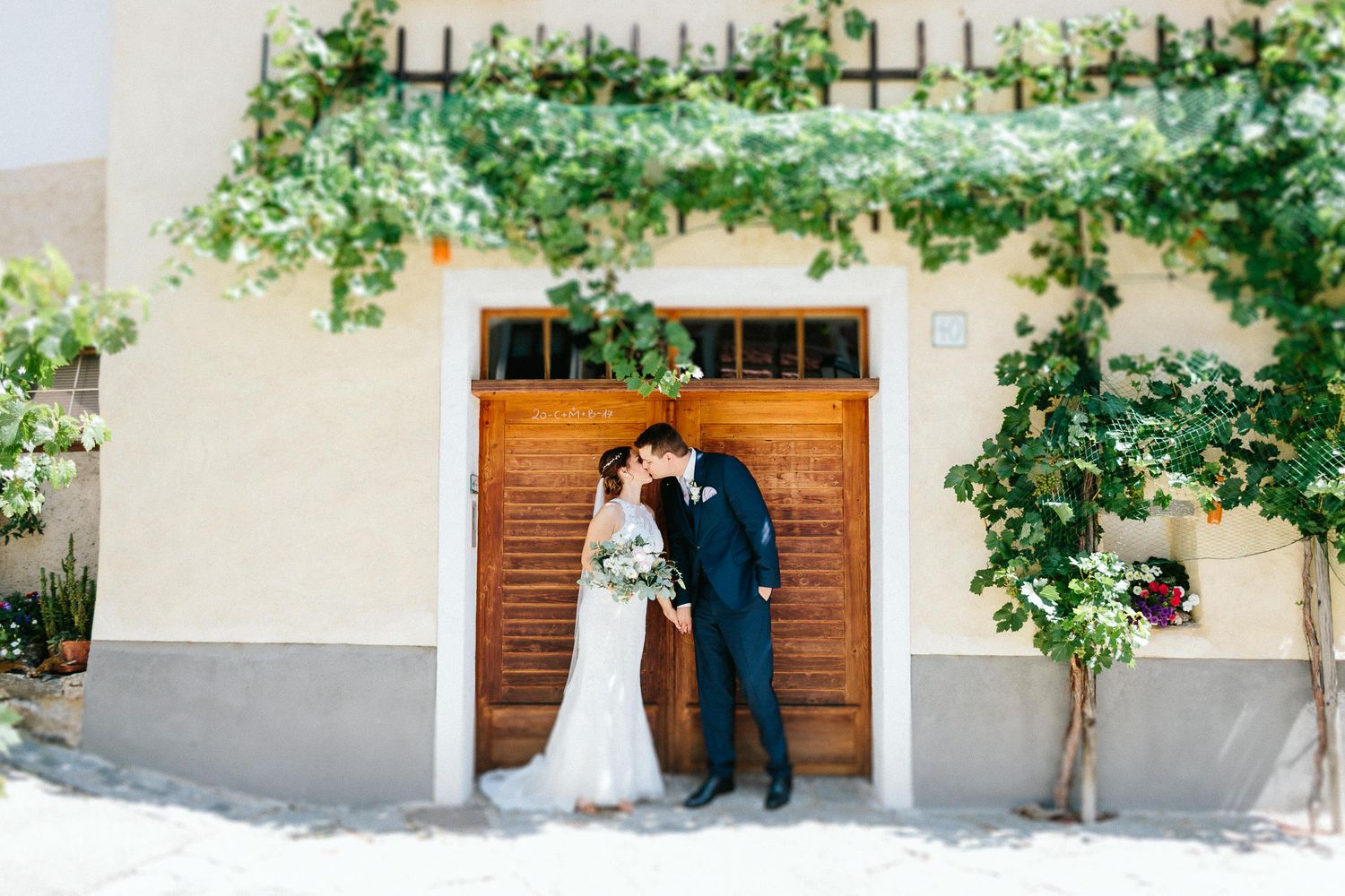 Bride and groom share a kiss in front of a rustic wooden door framed by lush green vines.