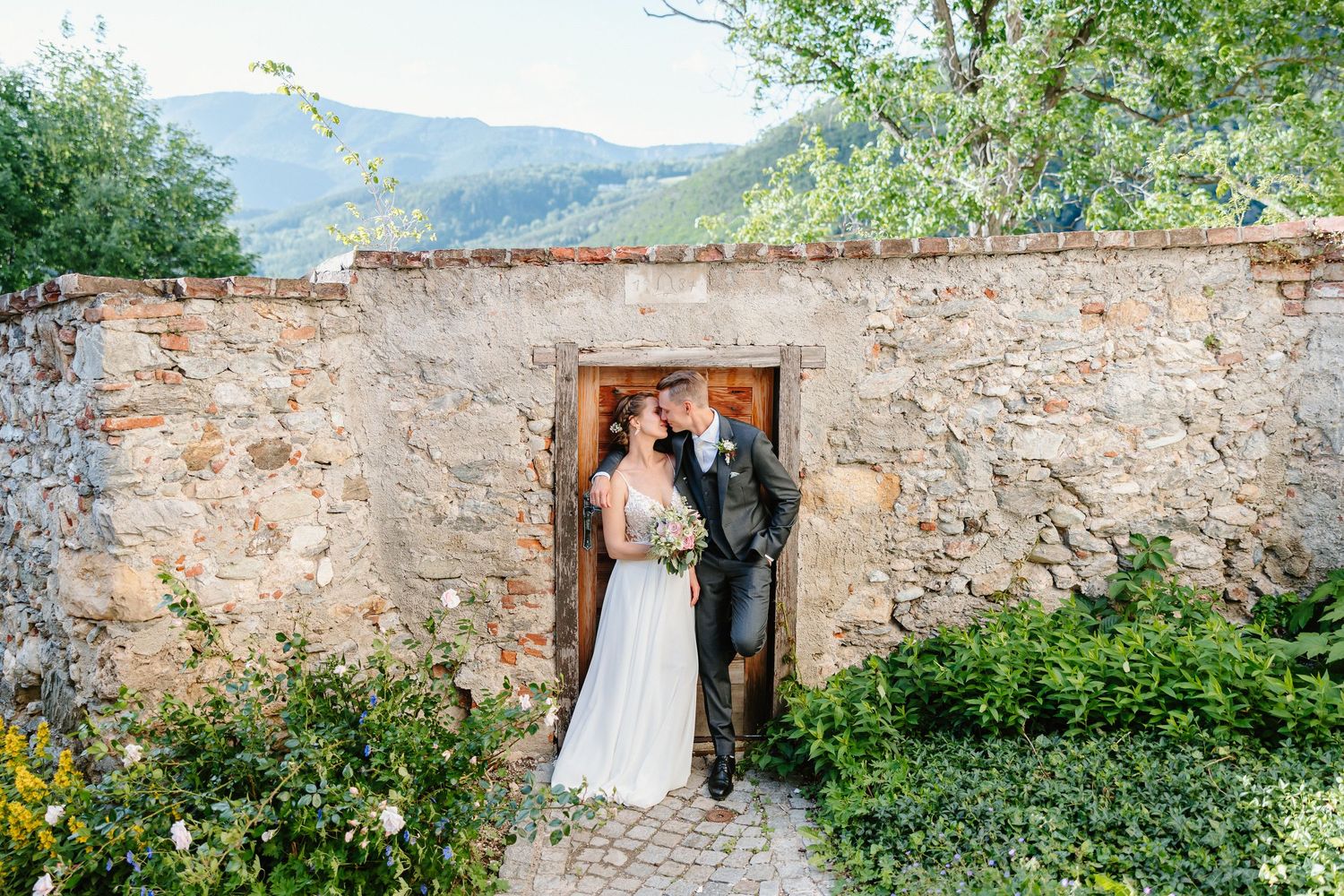 Bride and groom embracing in a charming stone archway doorway with scenic mountain landscape backdrop.
