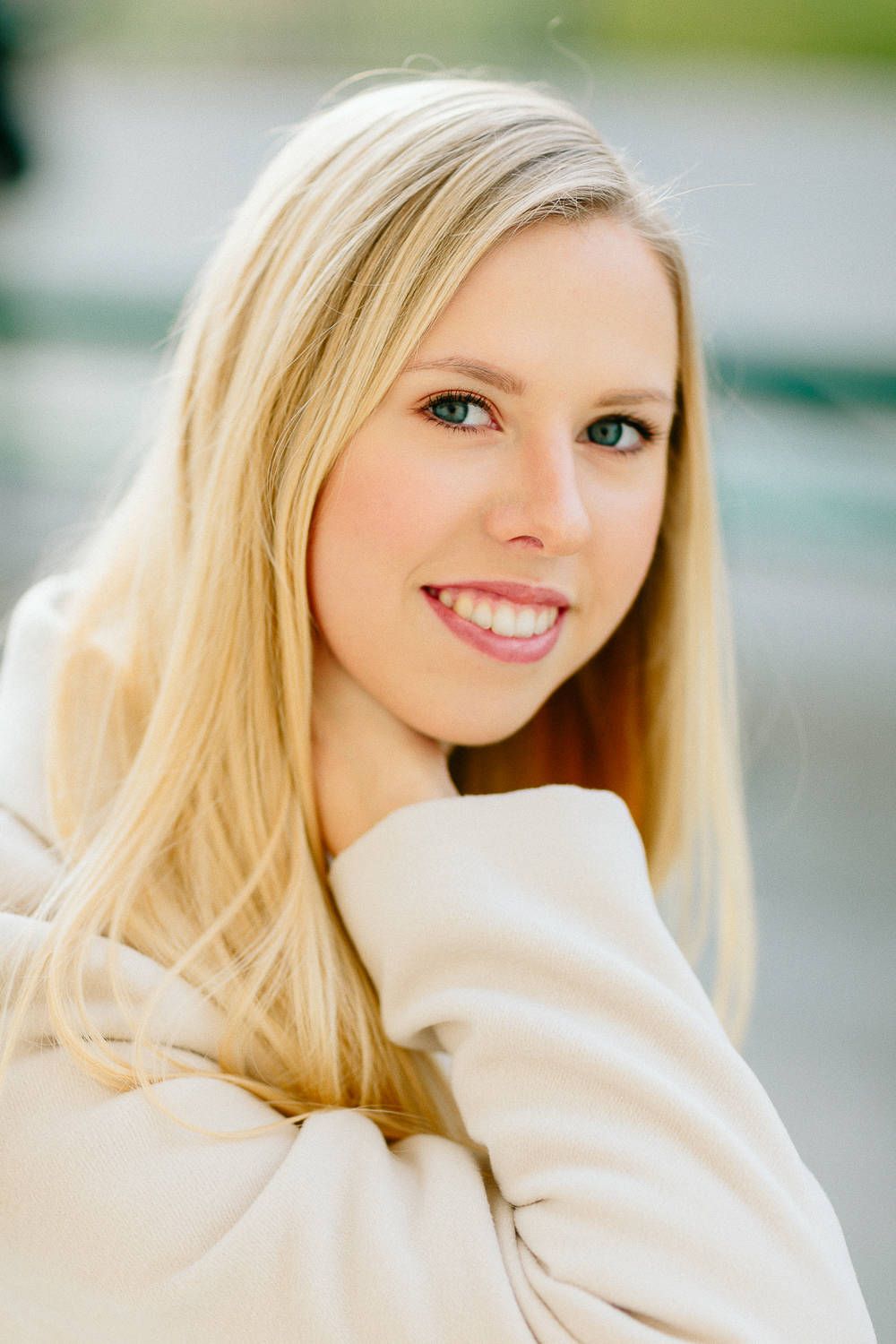 Young blonde woman with blue eyes smiling warmly in a cream sweater outdoors.