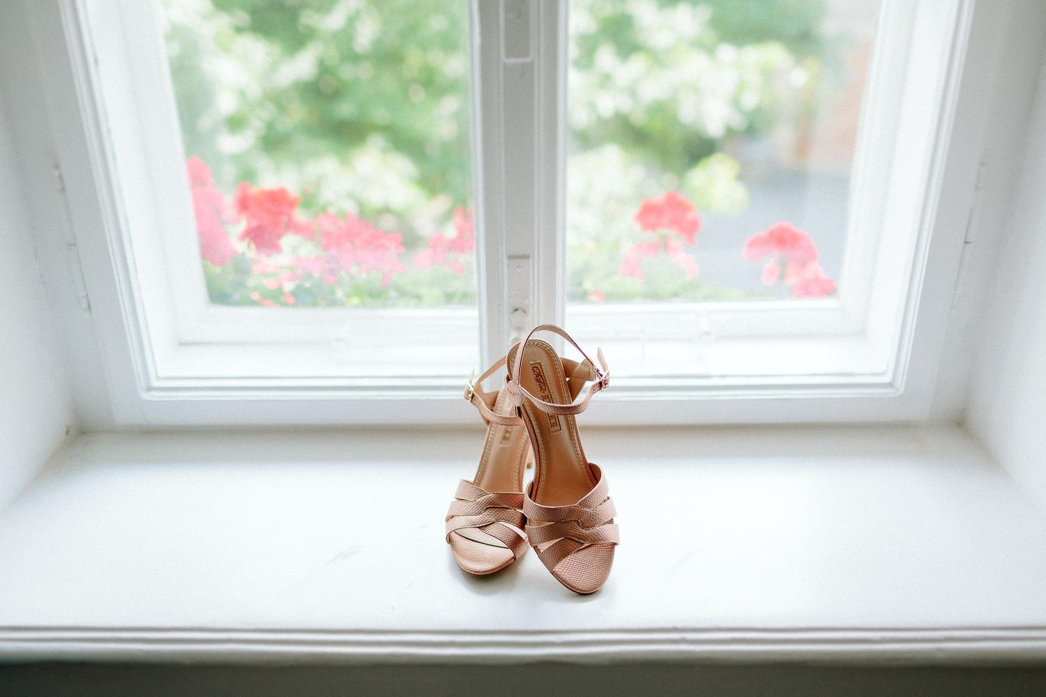 Elegant nude strappy bridal heels displayed on a white window sill with colorful flowers in background.