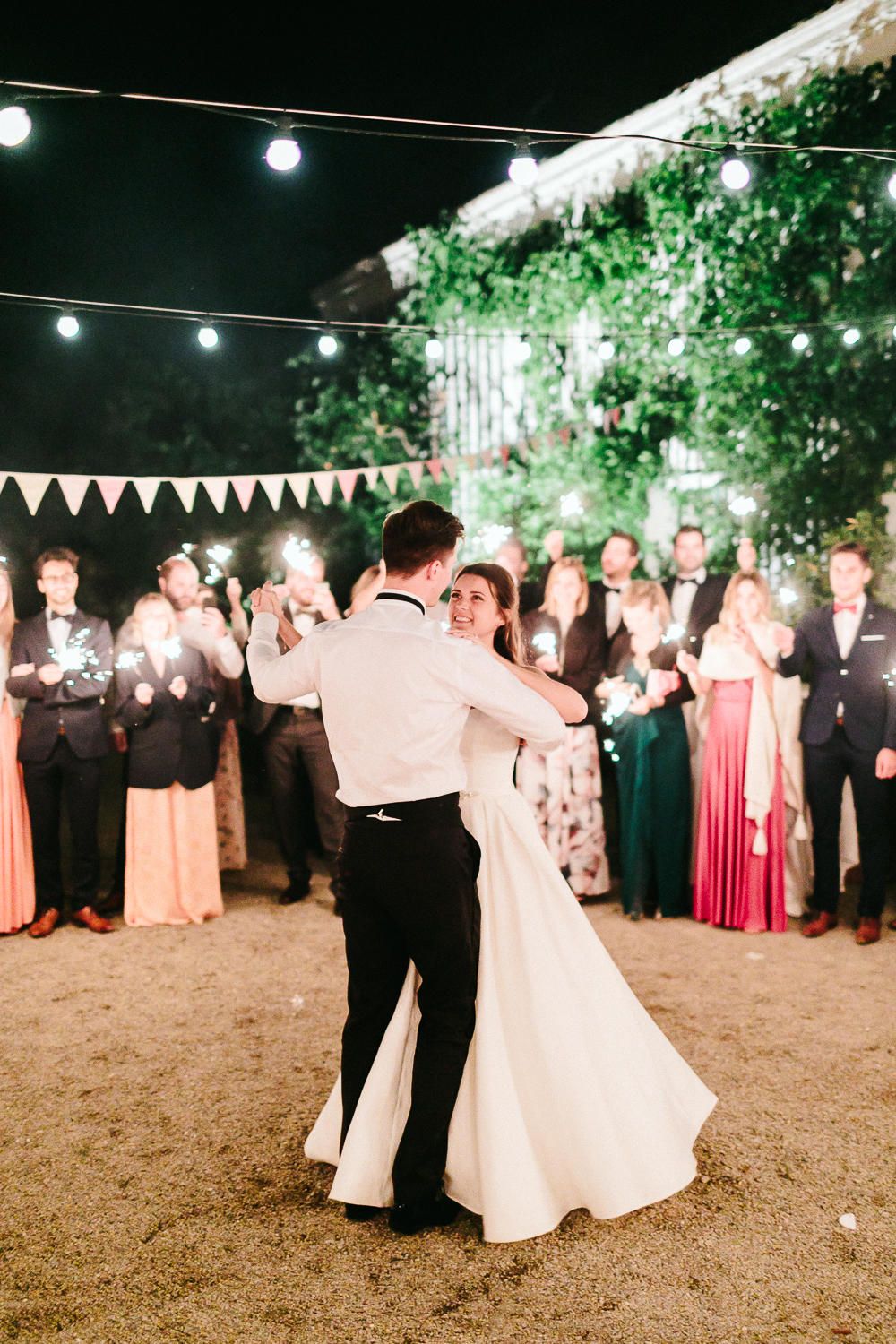 Bride and groom share their first dance outdoors at night surrounded by guests holding sparklers.