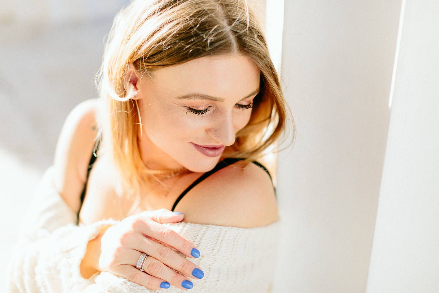 Young blonde woman in white off-shoulder sweater looking down thoughtfully with blue nail polish.