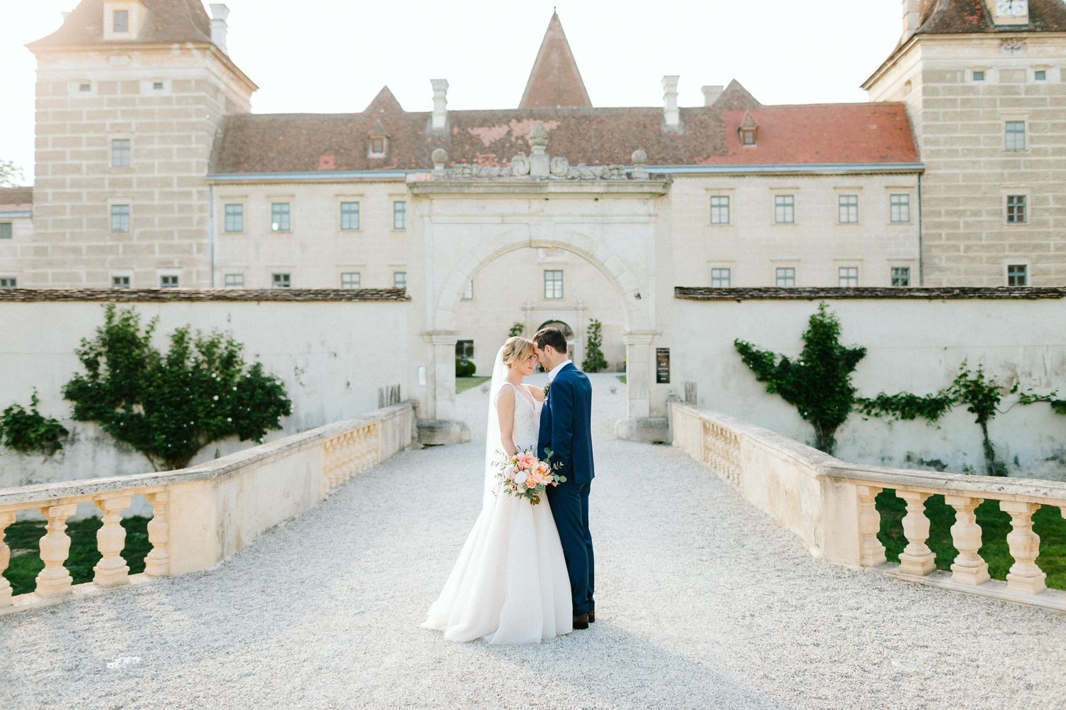Bride and groom share a romantic moment on a stone bridge outside an elegant European castle wedding venue.