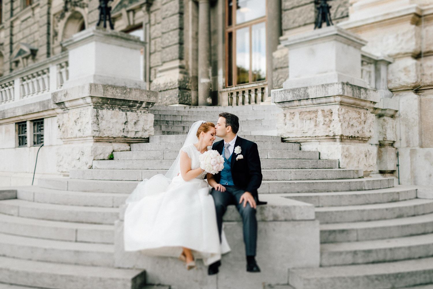 Bride and groom sitting closely on stone steps of a grand classical building sharing a romantic moment.