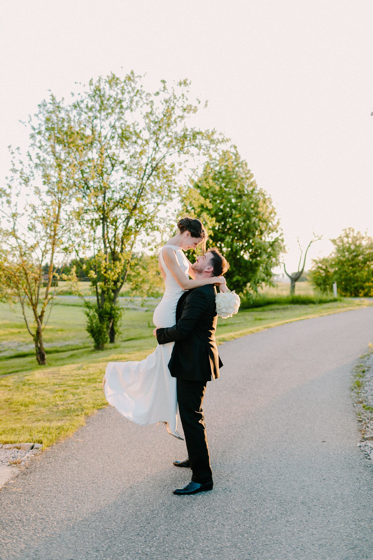 Groom lifts bride joyfully on a tree-lined road during golden hour wedding portraits.