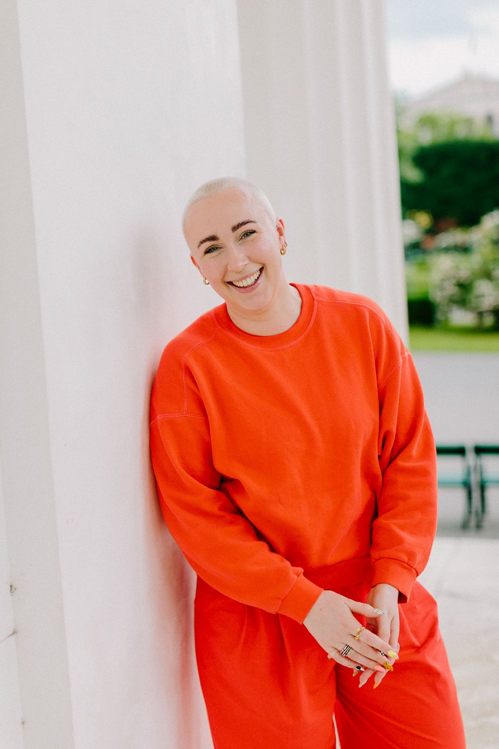 Confident bald woman in an orange sweatsuit smiling while leaning against a white pillar outdoors.