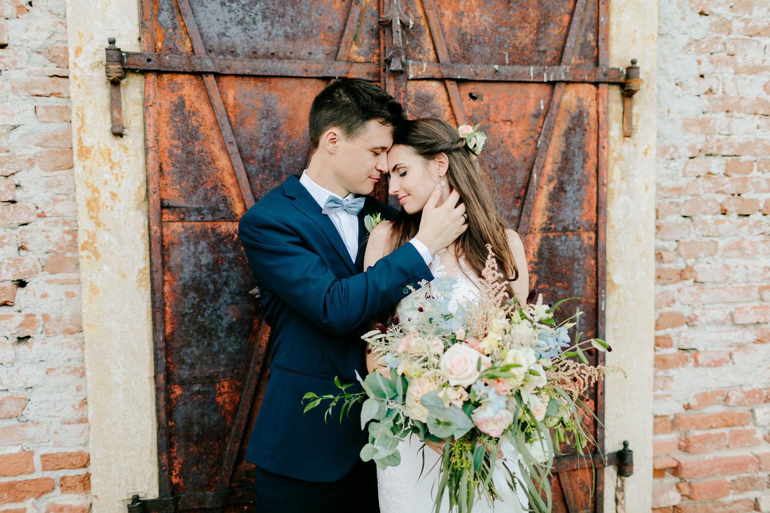 Bride and groom share an intimate moment in front of rustic wooden doors, bride holding a lush bouquet.