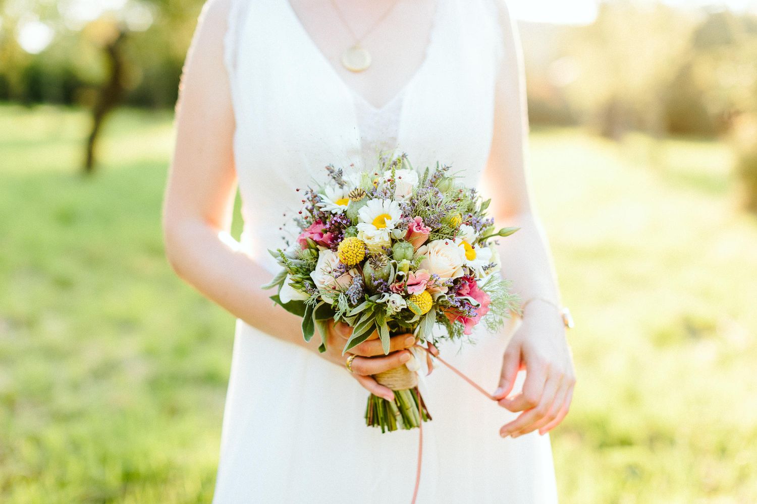 Bride in white sleeveless dress holding a colorful wildflower bouquet with pink and yellow blooms outdoors.