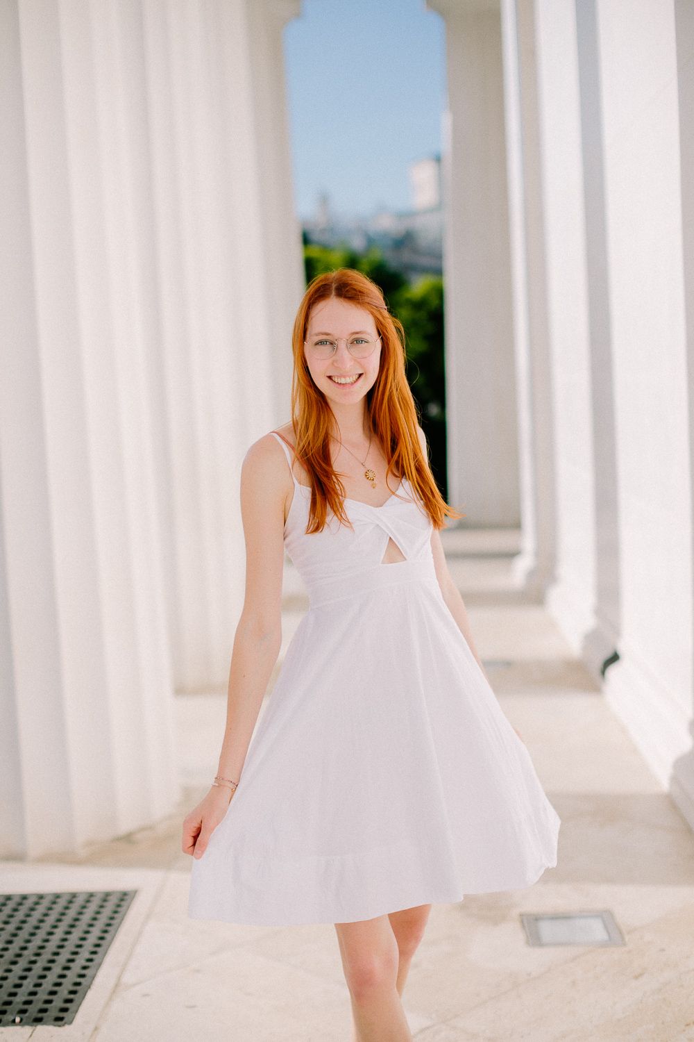 Young redhead woman in a white sundress smiling between white columns outdoors.