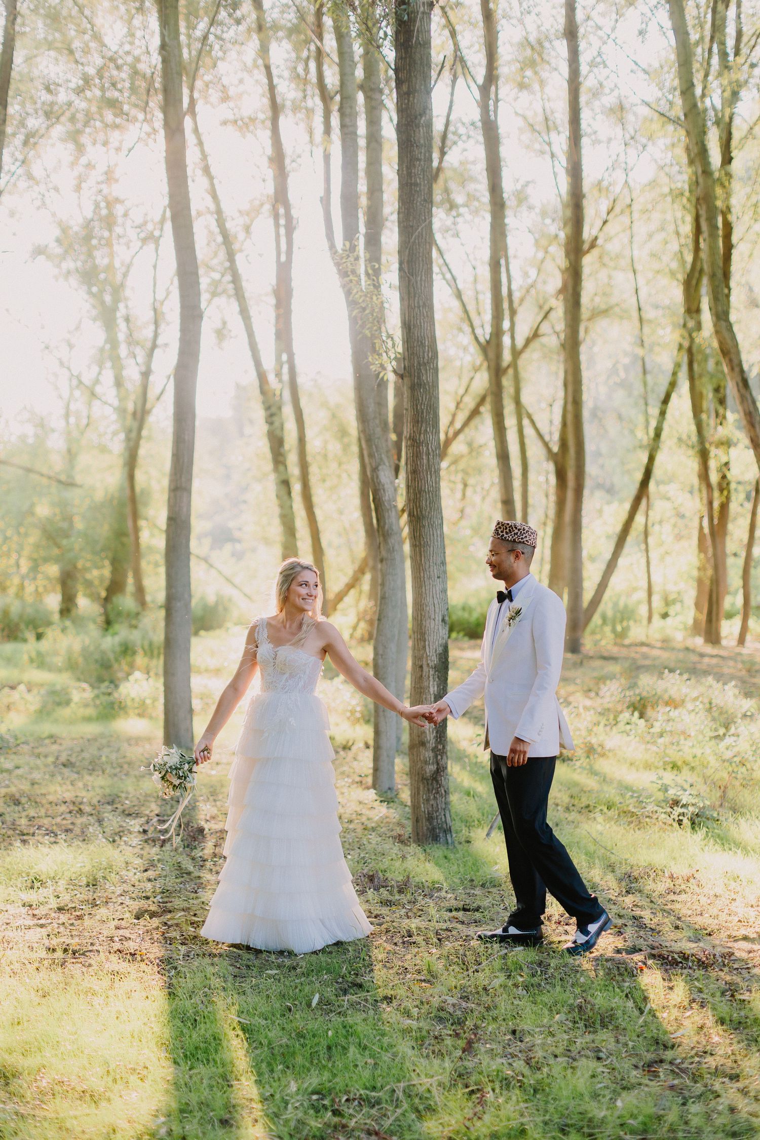Bride and groom holding hands walking through sunlit forest, groom wearing traditional patterned cap.