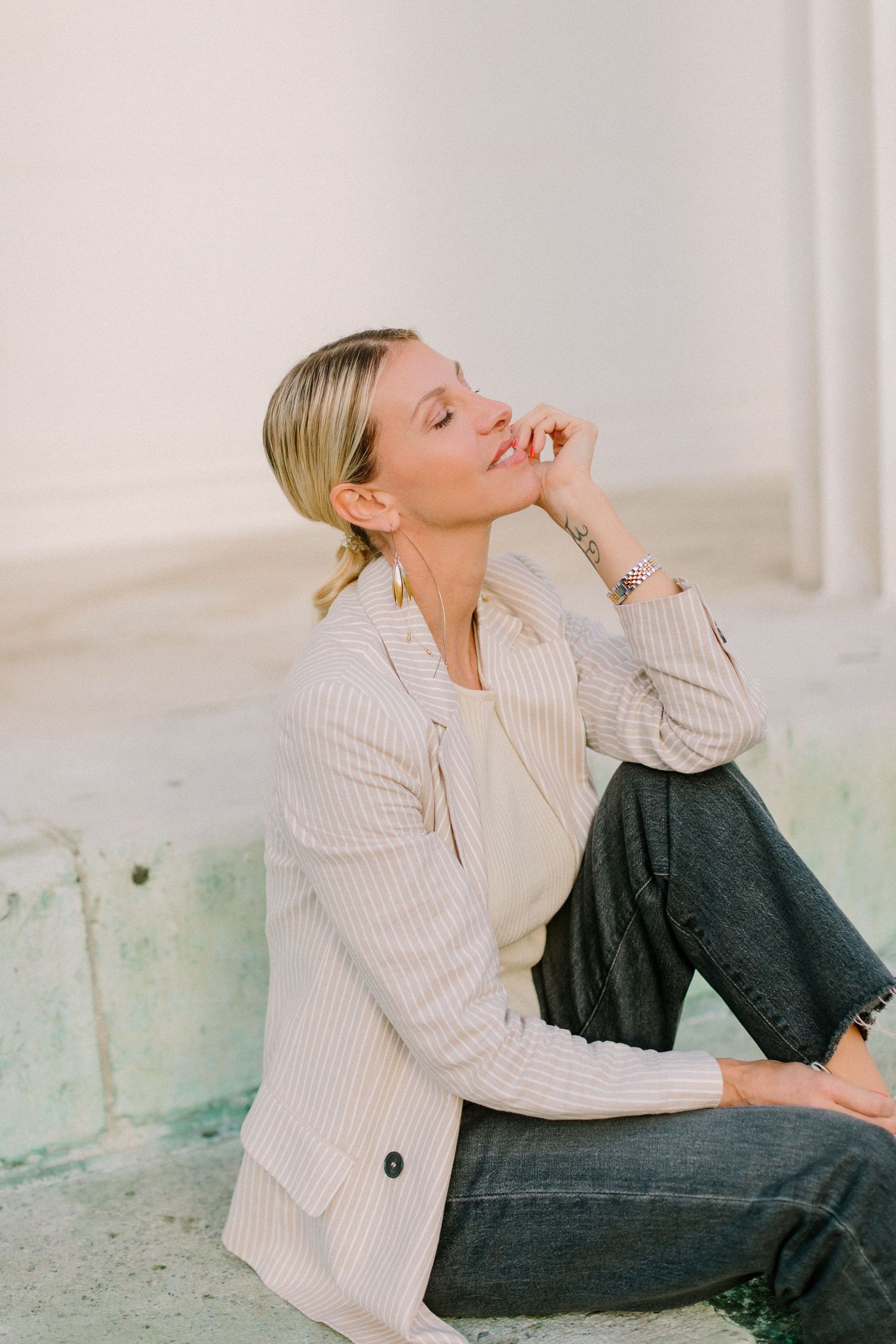 Blonde woman in striped blazer sits relaxed on steps in soft natural light.