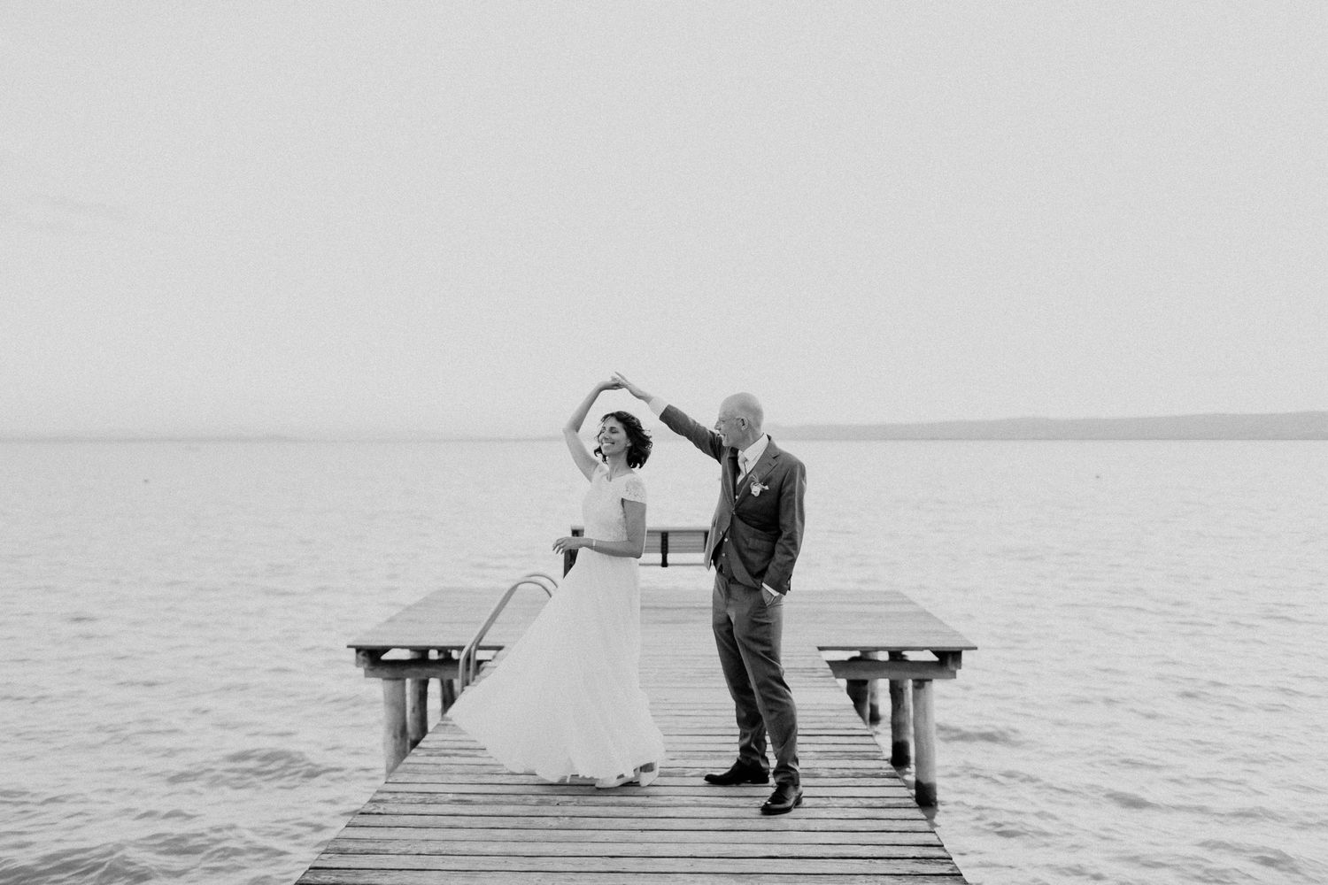 Black and white photo of bride and groom dancing on a wooden pier over calm lake.