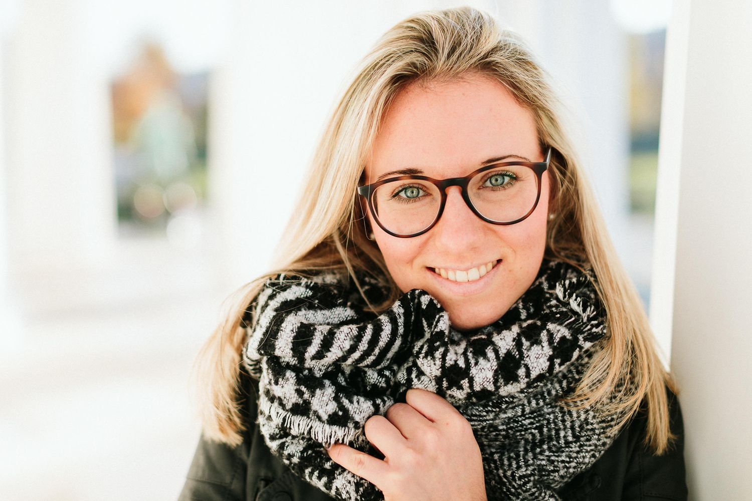 Smiling blonde woman wearing glasses and cozy knit scarf in bright outdoor portrait.