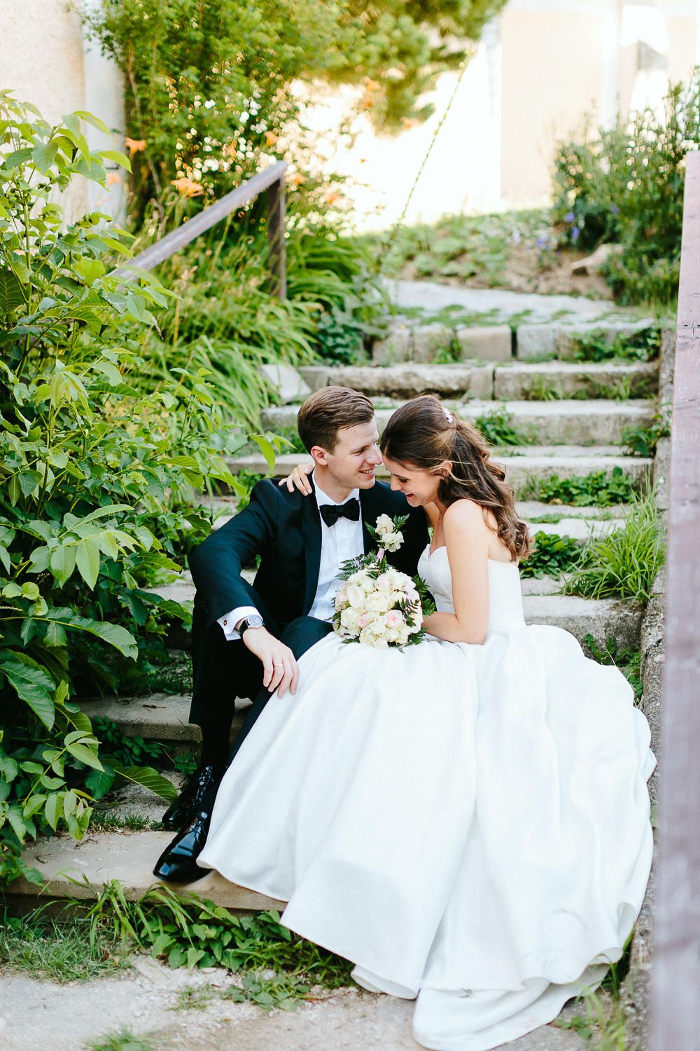 Bride and groom sitting on stone garden steps surrounded by green foliage sharing an intimate moment.