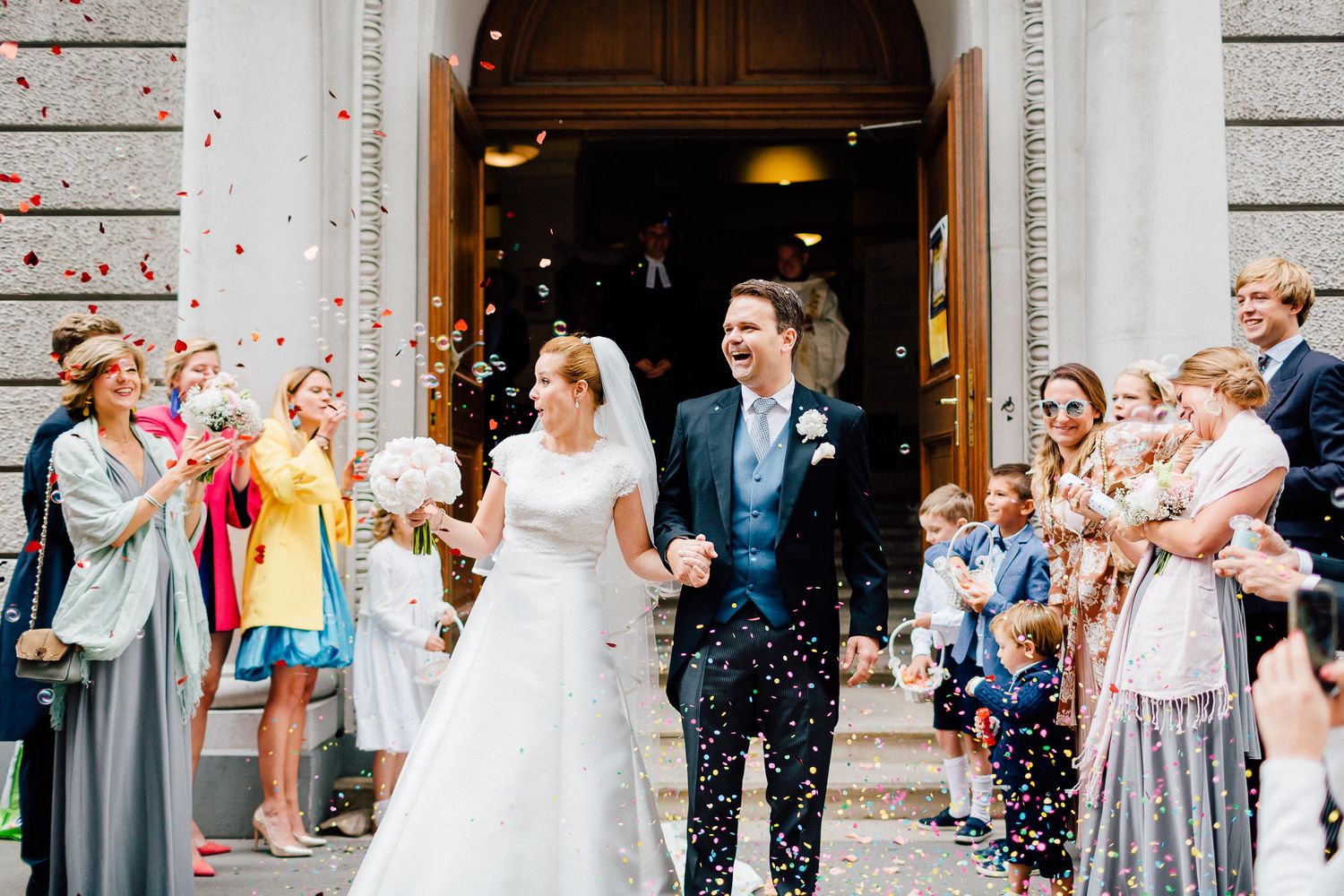 Joyful bride and groom exit church surrounded by guests throwing colorful confetti after ceremony.