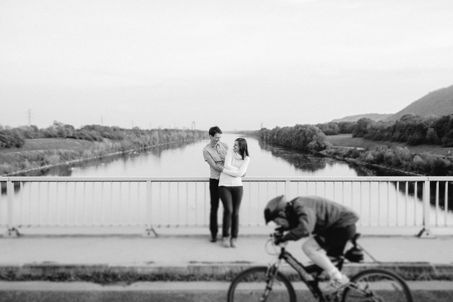 Couple sharing a romantic moment on a bridge over a calm river while a cyclist passes by.