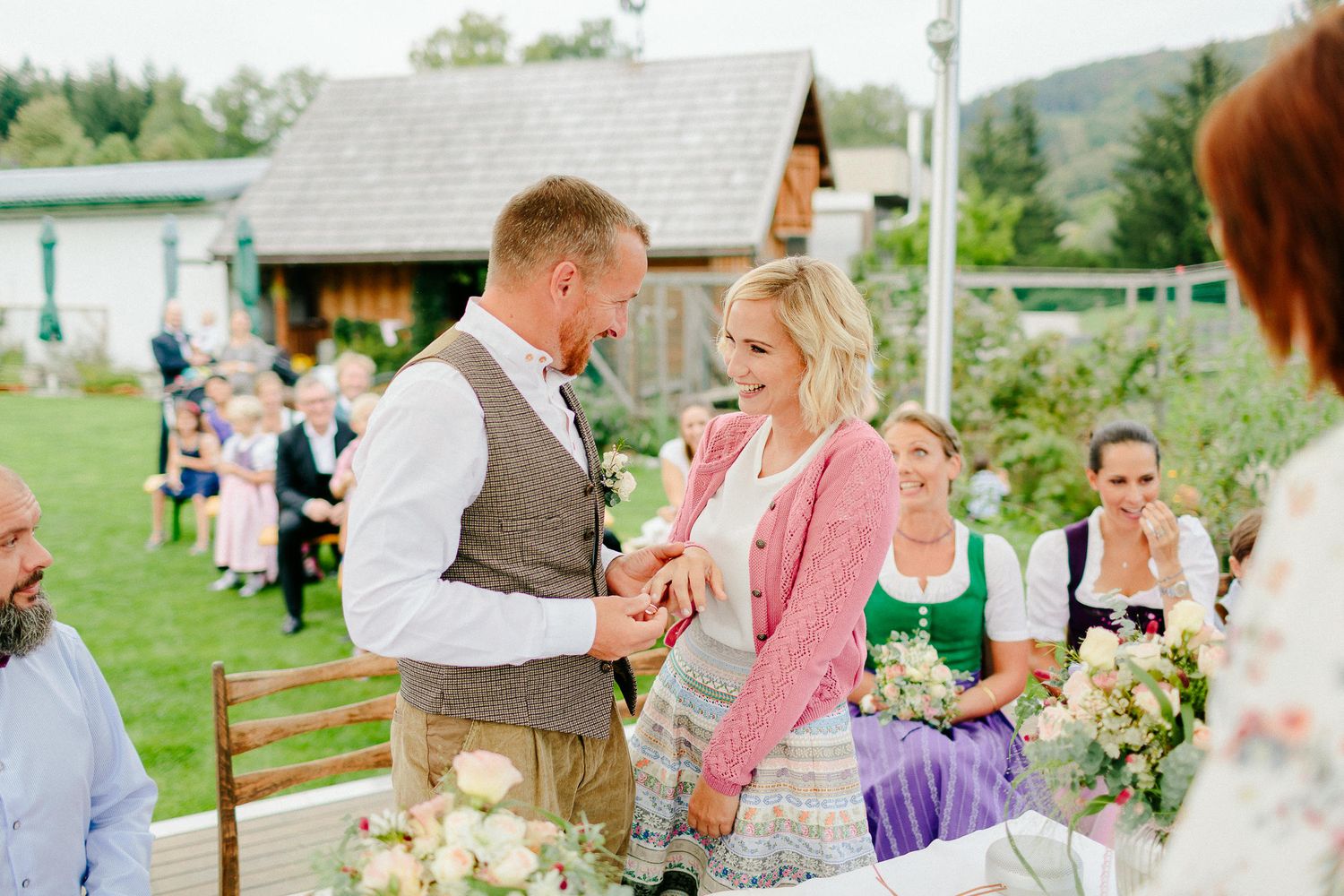 Groom places ring on bride's finger during outdoor wedding ceremony with guests in traditional attire.