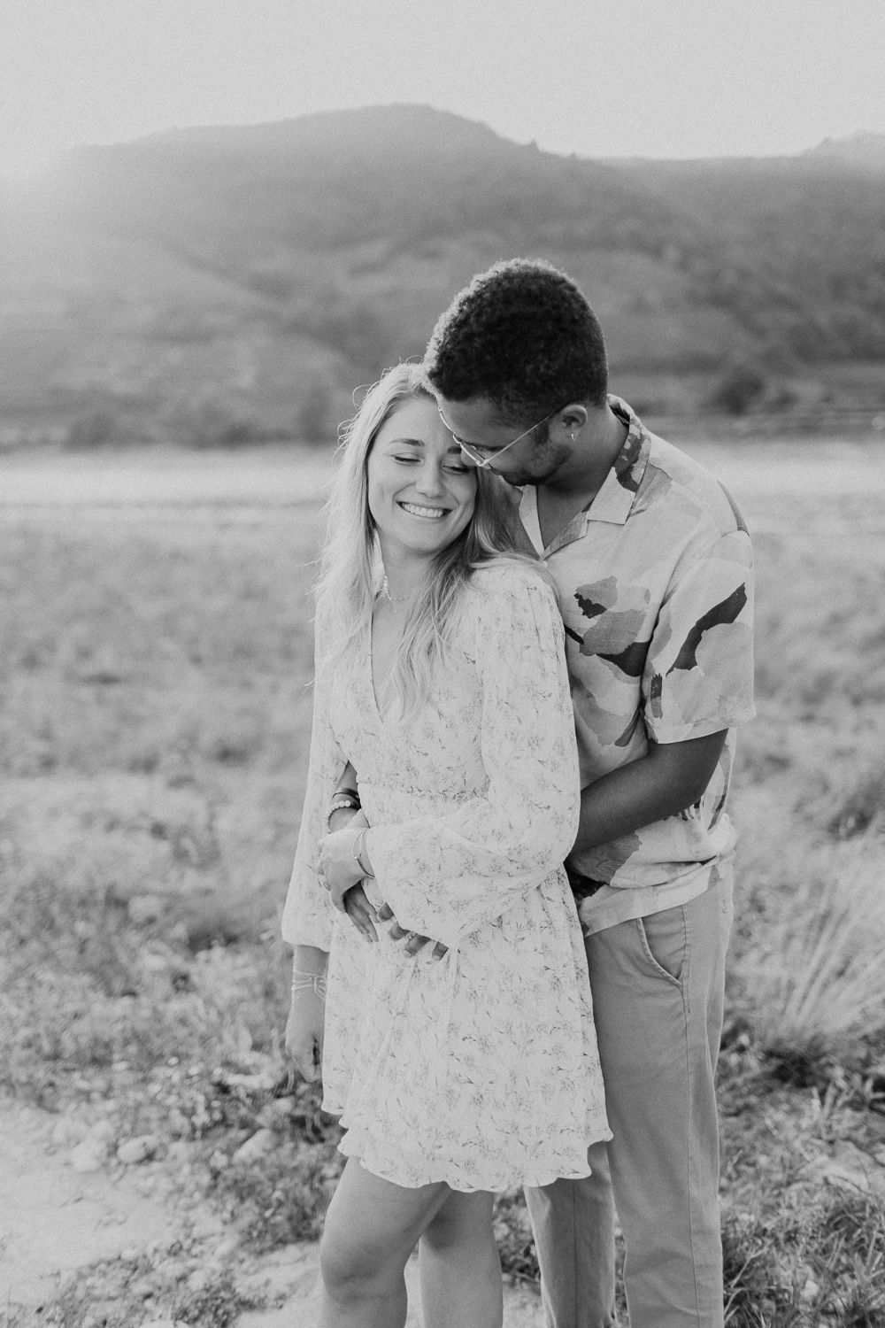 Interracial couple sharing a sweet kiss in a scenic field with rolling hills at sunset.
