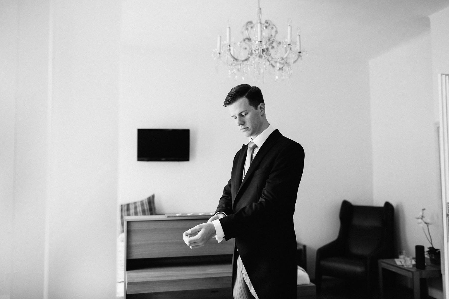 Black and white photo of groom in dark suit adjusting cufflinks in an elegant hotel room.