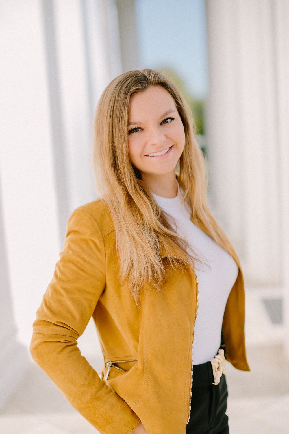 Smiling young woman in yellow suede jacket and white top posing between white columns.