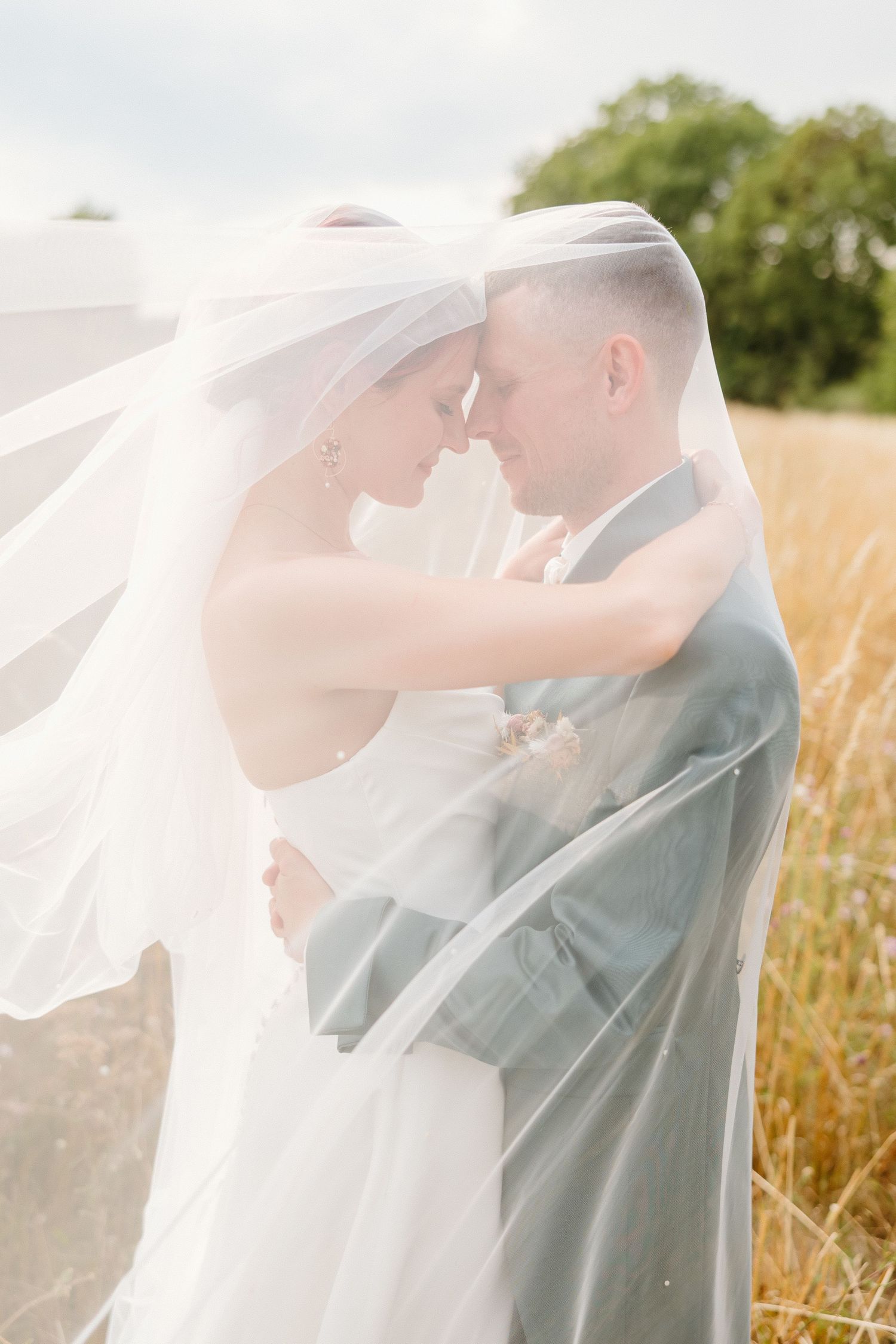 Bride and groom share intimate moment wrapped together under flowing white veil in field.