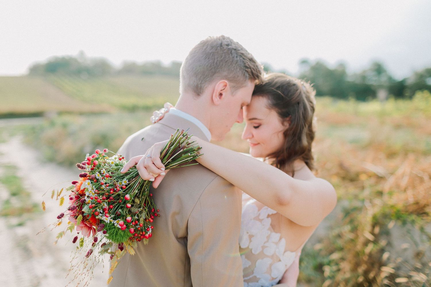 Couple embraces tenderly in a sunlit field, bride holding a vibrant red wildflower bouquet.