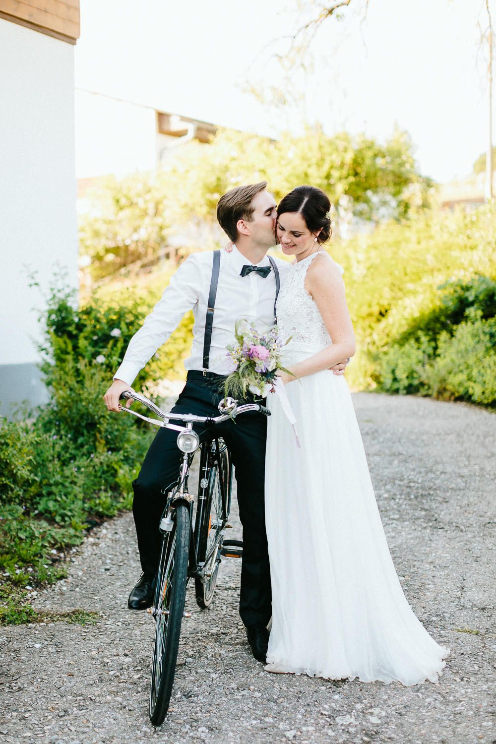 Groom in suspenders and bow tie kissing bride beside vintage bicycle in sunny outdoor garden setting.
