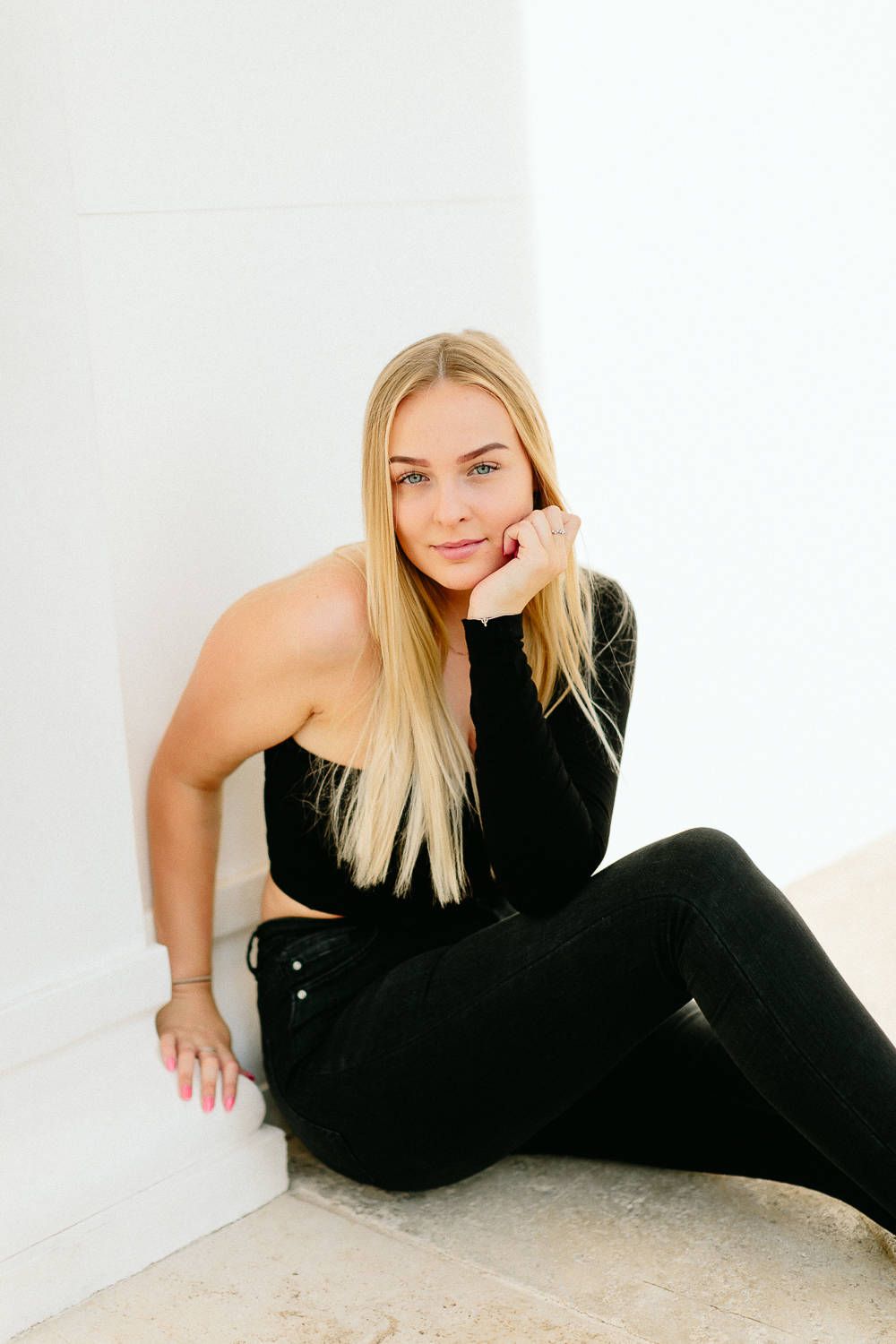 Blonde woman in all-black outfit sitting gracefully against a white wall on marble floor.