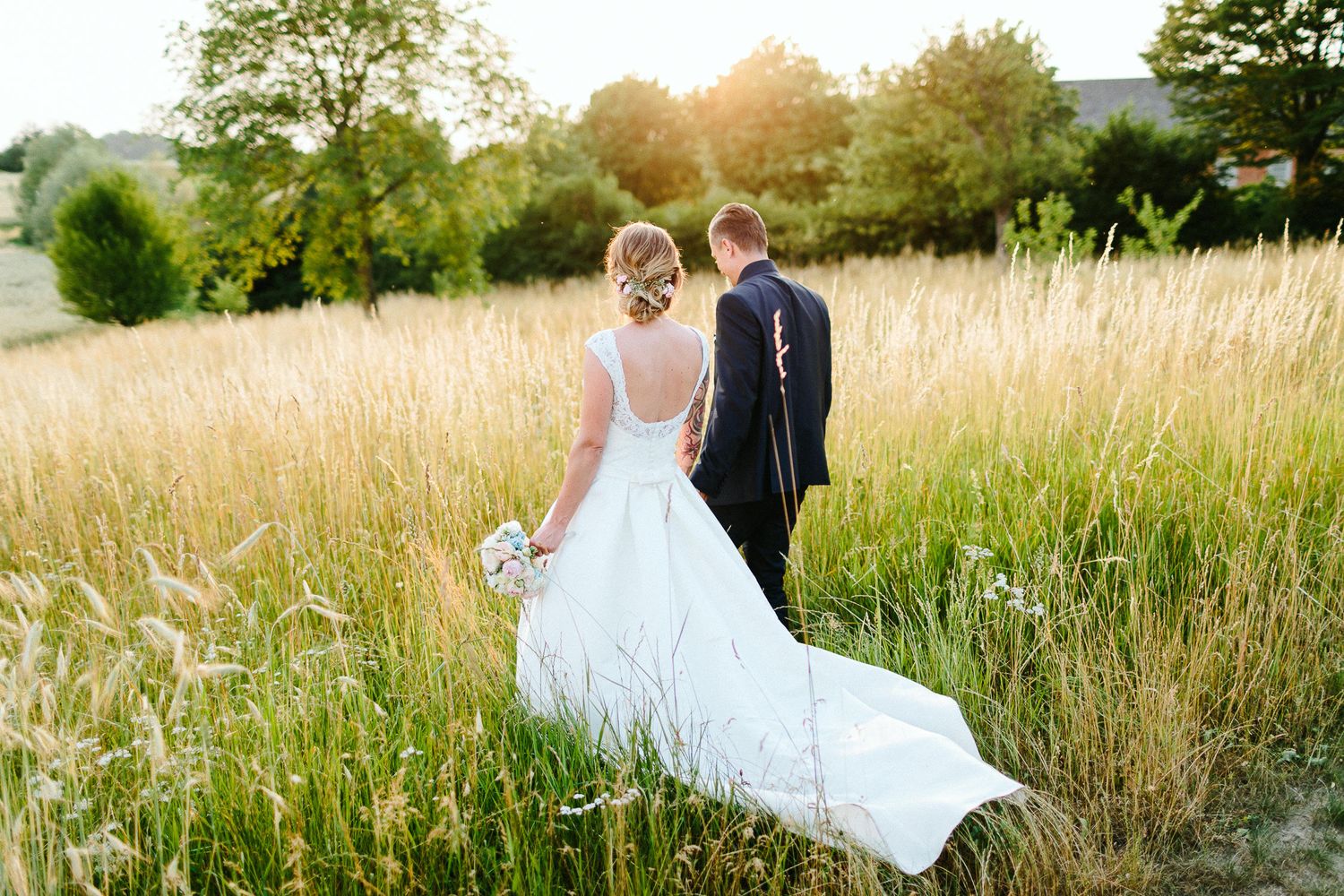 Wedding couple walks hand in hand through a golden field of tall grass at sunset.